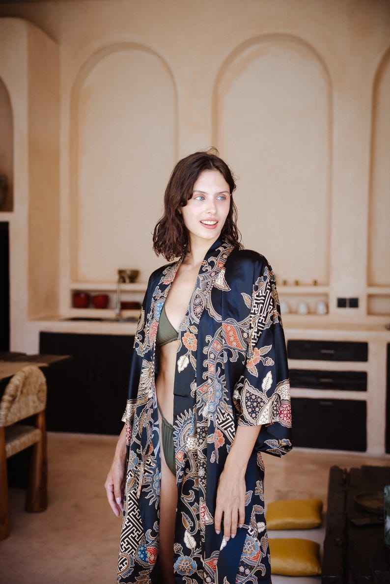 May include: A woman wearing a black silk robe with a colorful floral pattern. The robe has a shawl collar and long sleeves. The woman is standing in a room with a white wall and a wooden floor.