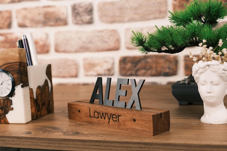 May include: A wooden desk nameplate with the name "ALEX" in black letters and the word "Lawyer" engraved below. The nameplate is on a wooden desk with a potted bonsai tree and a white bust in the background.