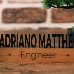 May include: Wooden desk nameplate with the name "ADRIANO MATTHEW" and the title "Engineer" engraved in black lettering.