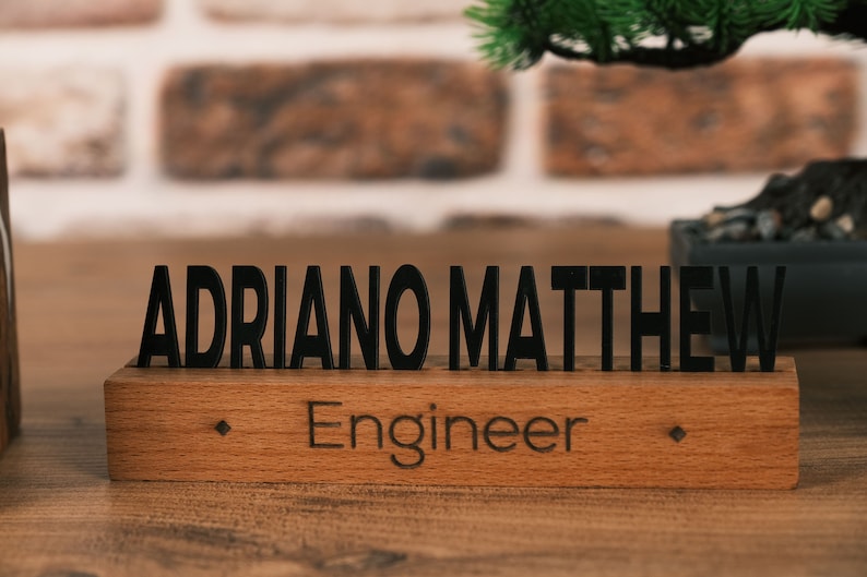 May include: Wooden desk nameplate with the name "ADRIANO MATTHEW" and the title "Engineer" engraved in black lettering.