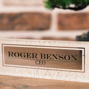 Op de afbeelding: Een heldere glazen naamplaat voor op het bureau met een bladgouden patroon en een rechthoekige gouden plaat met de tekst "ROGER BENSON CEO".