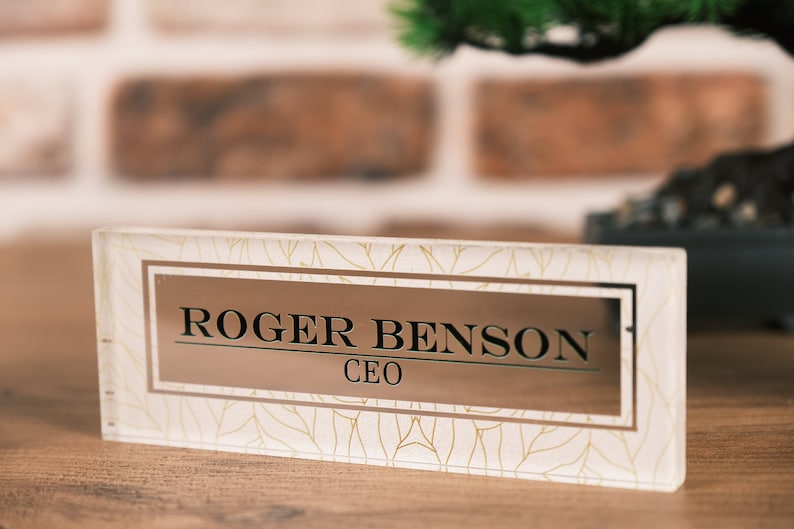 Op de afbeelding: Een heldere glazen naamplaat voor op het bureau met een bladgouden patroon en een rechthoekige gouden plaat met de tekst "ROGER BENSON CEO".