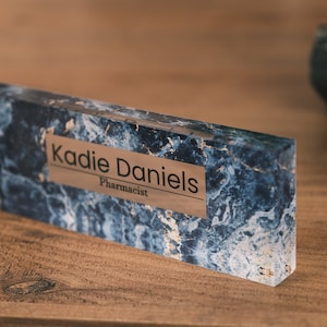 Op de afbeelding: Een blauwe en witte marmeren naamplaat voor op een bureau met een gouden plaat waarop "Kadie Daniels, Apotheker" staat.