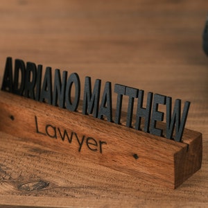 May include: A wooden desk nameplate with the name "ADRIANO MATTHEW" and the title "Lawyer" engraved in black lettering.