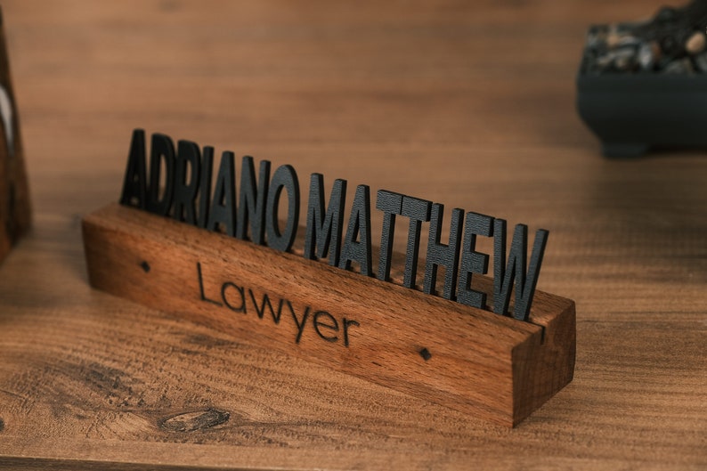 May include: A wooden desk nameplate with the name "ADRIANO MATTHEW" and the title "Lawyer" engraved in black lettering.