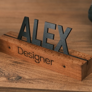May include: A wooden desk nameplate with the name "Alex" in black letters and the word "Designer" engraved below. The nameplate is on a wooden surface.