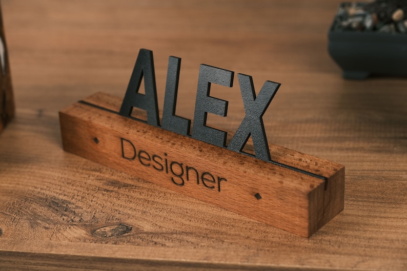 May include: A wooden desk nameplate with the name "Alex" in black letters and the word "Designer" engraved below. The nameplate is on a wooden surface.