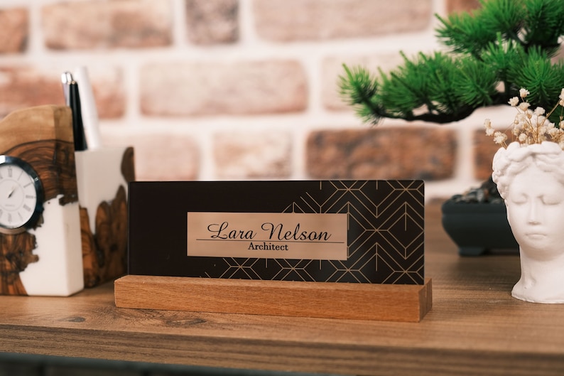 Op de afbeelding: Een houten bureau-naamplaatje met een zwarte achtergrond en een gouden geometrisch patroon. Het naamplaatje heeft een koperkleurig naamplaatje met de tekst "Lara Nelson Architect" erop.