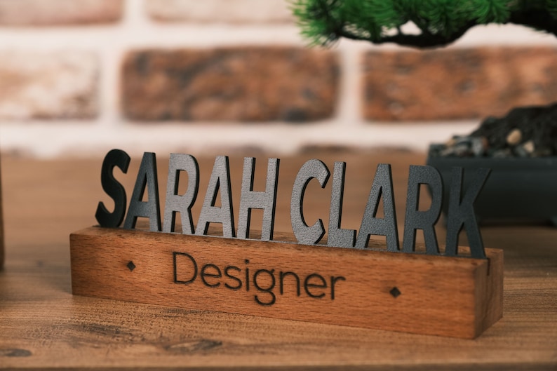 May include: A wooden desk nameplate with the name "SARAH CLARK" in black letters and the title "Designer" engraved below.