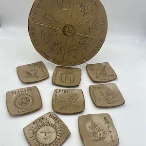 May include: A set of eight ceramic plates with Celtic symbols and runes. The plates are beige and feature various symbols, including a sun, a pumpkin, a holly sprig, a crescent moon, a triquetra, a lily, a flame, and a sheaf of wheat. Each plate has a different symbol and runes around the edge.