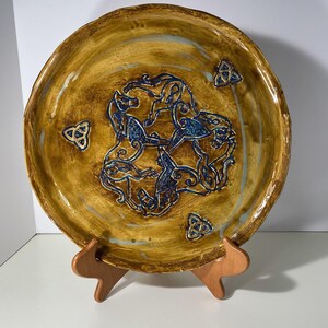 May include: A handcrafted ceramic plate with a brown and blue design. The plate features a central motif of stylized horses in blue, surrounded by Celtic knot symbols. The plate is displayed on a wooden stand.