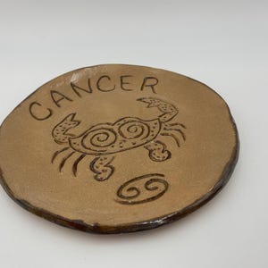 May include: A round, tan ceramic plate with the word "CANCER" etched at the top. A crab illustration is in the center, with the Cancer symbol below. The plate has a dark brown rim.