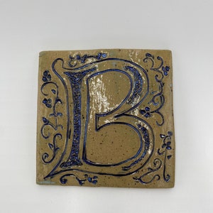 May include: A ceramic tile with a blue and white glazed finish featuring a decorative letter "B" in a stylized font. The letter is surrounded by a floral design.