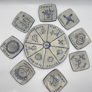 May include: A set of ten ceramic tiles with black and white designs. The tiles feature various symbols and runes, including a cauldron, a tree, a sun, and a moon. The tiles are arranged in a circle, with a central tile featuring a spiral design.