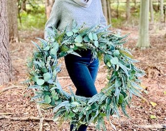 May include: A large, circular wreath composed of various artificial green foliage, including eucalyptus leaves, ferns, and small sprigs. The wreath features a dense arrangement of layered leaves in shades of sage and forest green, creating a full, textured appearance.