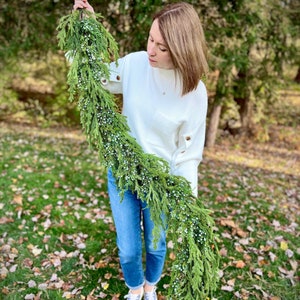 May include: A long, green garland with small white berries is held up. The garland is made of evergreen branches. The person is wearing a white sweater, blue jeans, and sneakers. The background is a grassy area with trees.