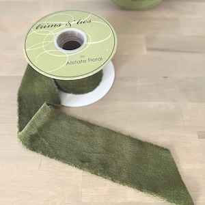 May include: A spool of olive green velvet ribbon with the text "trims & ties" and "by Allstate Floral" printed on the label. The ribbon is unrolled, showing its textured surface.