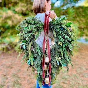 May include: A fresh evergreen wreath with pinecones and sprigs of greenery. The wreath is adorned with a deep red ribbon and hanging gold bells. The wreath is being held up by a person wearing a gray sweater and blue jeans.