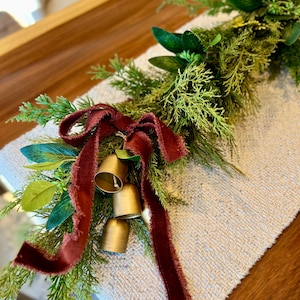 May include: A festive Christmas garland featuring faux greenery, including pine and leafy sprigs. The garland is adorned with a burgundy velvet ribbon and three gold bells, creating a warm, inviting holiday decoration.