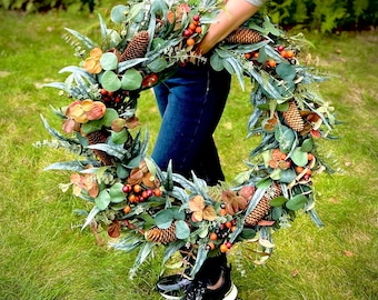 May include: A decorative wreath featuring a mix of greenery, orange berries, and pinecones. The wreath has a circular shape and is held by a person wearing a gray sweater and blue jeans. The wreath is set against a backdrop of green grass.