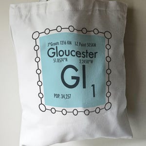 May include: A white tote bag with a blue and black graphic design. The graphic features the text "Gloucester" with coordinates and a stylized chemical element symbol "GI" with the number "1" and the text "POP. 34,257".