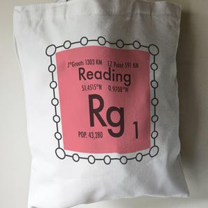 May include: White tote bag featuring a pink and black graphic of a periodic table element. The element is labelled "Rg" for the town of Reading, England. The graphic also includes the town's coordinates and population.