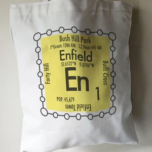 May include: A white tote bag with a yellow and black graphic design. The graphic is a stylized representation of a tube map with the text "Enfield" and "En 1" in black. The graphic also includes the text "Bush Hill Park", "J°Groats 1286 KM", "LZ Point 697 KM", "51.6532°N 0.0706°W", "Forty Hill", "Bulls Cross", "Enfield Town", and "POP. 45,679".