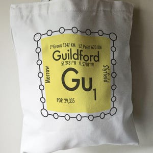 May include: A white tote bag with a yellow and black graphic design. The graphic features a stylized map of Guildford, England, with the text "Guildford", "51.2431°N 0.5701°W", "Gu1", and "POP. 29,335".