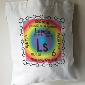 May include: A white tote bag with a colourful graphic design featuring the word "Leeds" and the letters "Ls" in a rainbow circle. The graphic also includes the coordinates "53.8185°N 1.5663°W" and the text "POP. 47,533" and "6". The graphic is surrounded by a black circle with smaller circles around it.