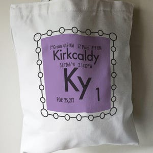 May include: A purple tote bag with a white design that looks like a periodic table element. The element is labelled "Ky" and has the text "Kirkcaldy" above it. The text "J°Groats 449 KM LZ Point 1119 KM" is above the element name. The text "56.1266°N 3.1412°W" is below the element name. The text "POP. 25,212" is below the element symbol.
