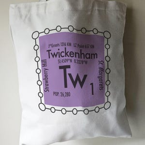 May include: A white tote bag with a purple graphic displaying the name "Twickenham" and related information. The bag has a handle and is made of a light-colored fabric, perfect for carrying items.
