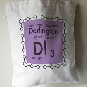 May include: A purple and white tote bag with a graphic design of a city name, coordinates, and population. The graphic is surrounded by a black circle with smaller circles around it. The text reads "Darlington, 54.5312°N 1.5690°W, DI 3, POP. 44,095".