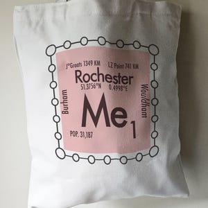 May include: A white tote bag with a pink and black design. The design features a circle with the text "Rochester" and the coordinates "51.3756°N 0.4998°E". The text "Me1" is also included in the design.