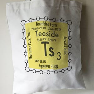 May include: A white tote bag with a yellow and black design. The design features the text "Teeside" with the coordinates "54.5619°N 1.1985°W" and the text "Ts3" with the population "POP. 29,393".