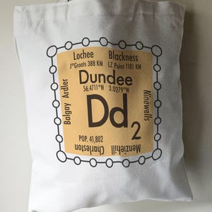 May include: A white tote bag with a brown and black graphic design. The graphic features a stylised map of Dundee, Scotland, with the city's name, coordinates, and population. The graphic also includes the text "Dd2" and the names of nearby towns.