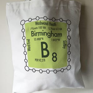 May include: A white tote bag with a green and black graphic design. The graphic features the text "Birmingham" with coordinates and the text "B8" with the population of 42,278. The graphic is surrounded by a black circle with smaller circles around it.