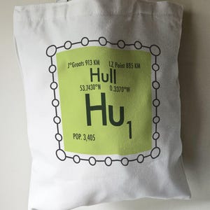May include: A white tote bag featuring a green and black graphic of the city name "Hull", styled to resemble a periodic table element. The graphic includes the city's coordinates, "53.7430°N 0.3370°W", and population, "POP. 3,405".