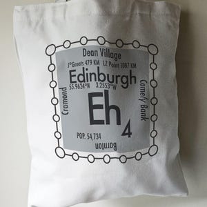 May include: White tote bag with a black and grey design featuring the text "Edinburgh Eh4" and the coordinates "55.9624°N 3.2553°W". The design also includes the names of nearby locations, such as "Dean Village", "Cramond", and "Comely Bank".