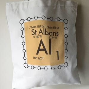 May include: A white tote bag featuring a brown and black graphic of a periodic table element. The element is labelled "Al" for Aluminium and has the number "1" below it. The graphic also includes the text "St Albans" with the coordinates "51.7487°N 0.3194°W" and the population "POP. 36,291".