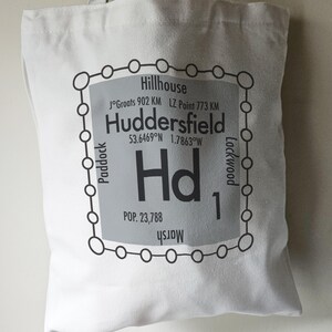May include: A white tote bag with a black and grey design. The design features a square with rounded corners and the text "Huddersfield" with the coordinates "53.6469°N 1.7863°W" and "POP. 23,788". The square is surrounded by a circle of smaller circles with the names of surrounding towns.
