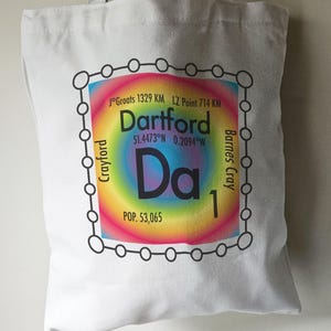 May include: White tote bag featuring a black and white graphic of a rainbow coloured circle with the text "Dartford" and "Da1" inside the circle. The text "JoGroats 1329 KM, LZ Point 714 KM" is above the circle. The text "51.4473°N 0.2094°W" is below the circle. The text "POP. 53,065" is below the circle. The text "Barnes Cray" and "Crayford" are on the right and left sides of the circle, respectively.