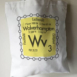 May include: A white tote bag with a yellow and black graphic design. The graphic features the text "Castlecroft", "J°Groats 1088 KM LZ Point 601 KM", "Wolverhampton", "52.5808°N 2.1483°W", "Wv", "POP. 29,731", and "3". The graphic also includes the text "Finchfield" and "Compton" written vertically on either side of the graphic.