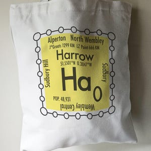 May include: A white tote bag featuring a yellow graphic of a tube station sign. The sign reads "Harrow" with the coordinates "51.5501°N 0.3047°W" and the population "POP. 48,931". The sign is surrounded by a black circle with smaller circles around it.