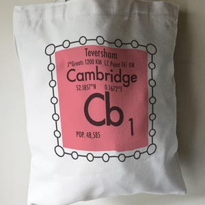 May include: A pink and white tote bag with a black outline of a periodic table element. The element is labelled "Cb" with the number "1" and the text "Cambridge" above it. The text "Teversham" is above the element name, and the text "J°Groats 1200 KM LZ. Point 761 KM" is below the element name. The text "52.1857°N 0.1672°E" is below the element number. The text "POP. 48,585" is below the element symbol.