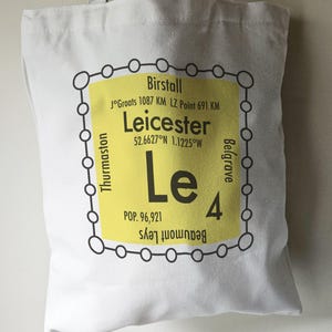 May include: A white tote bag with a yellow and black graphic design. The graphic features the text "Leicester" with the coordinates "52.6627°N 1.1225°W" and the text "Le 4" with the text "POP. 96,921" below. The graphic is surrounded by a black circle with smaller circles around the perimeter.