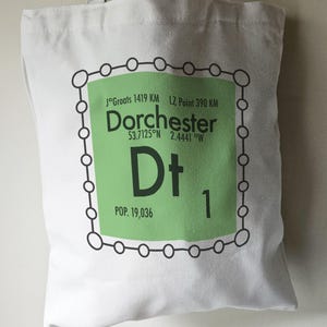 May include: White tote bag with a green and black graphic of a town square. The graphic includes the text "Dorchester", "53.7125°N 2.4441°W", "Dt", "POP. 19,036", and "1".