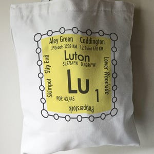 May include: A white tote bag featuring a yellow graphic of a train station stop sign. The sign reads "Luton" with the coordinates 51.8764°N 0.4246°W and a population of 43,445. The sign also lists nearby stops: Aley Green, Caddington, Skimpot, Slip End, Lower Woodside, and Pepperstock.