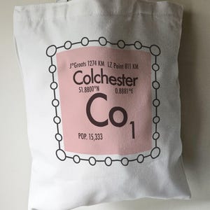 May include: A white tote bag featuring a pink and brown graphic of a periodic table element. The element is labelled "Co" for Colchester, with the city's coordinates and population listed below. The graphic is surrounded by a black circle with smaller circles around the perimeter.