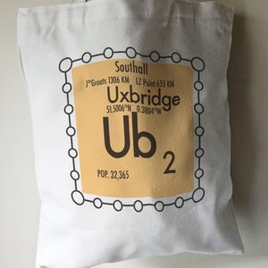 May include: A white tote bag with a black and orange graphic design. The graphic features a circle with the text "Southall", "Uxbridge", "51.5006°N 0.3804°W", "Ub2", and "POP. 32,365".