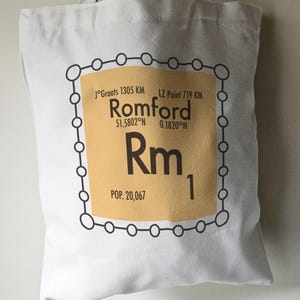 May include: A white tote bag featuring a brown and black graphic of a periodic table element. The element is labelled "Rm" for Romford, with the coordinates 51.5802°N 0.1820°W and a population of 20,067.